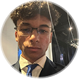 Imagem de Vitor Carneiro com expressão neutra, de oculos, sem barba, com apenas a parte superior do terno próxima ao pescoço amostra em um fundo iluminado.